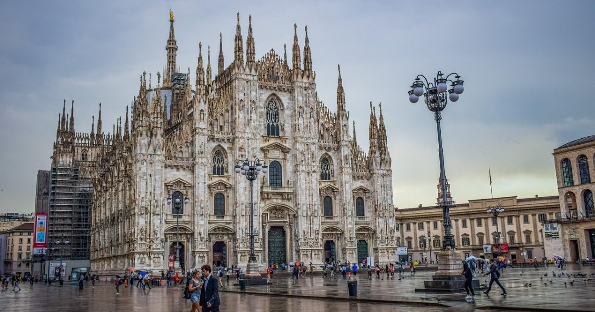 清晨的米兰大教堂广场上，一位背着双肩包的中国游客举起手机查看 milano italy App 上的步行导航路线，白鸽从身旁掠过，哥特式尖顶在晨光里泛着暖金色